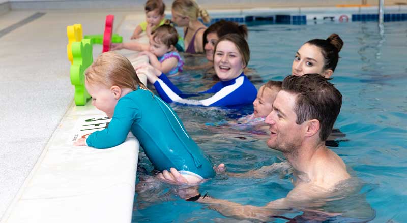 Toddlers holding onto the edge of a swimming pool with the support of adults