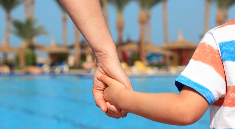 A mother guides her child into a pool area while holding their hand, showing proper parental supervision around water