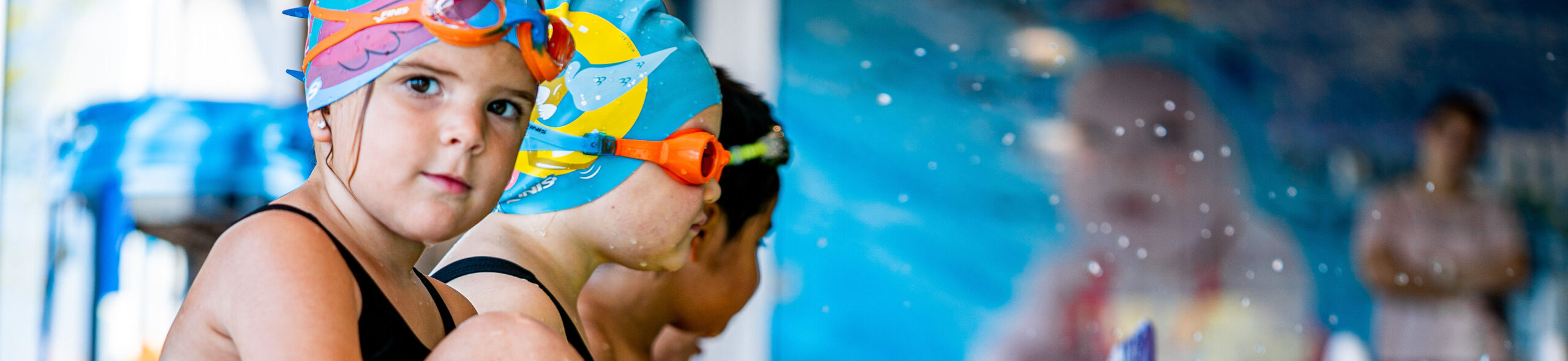 Children smiling during a swim lesson with an instructor in an indoor swimming pool.
