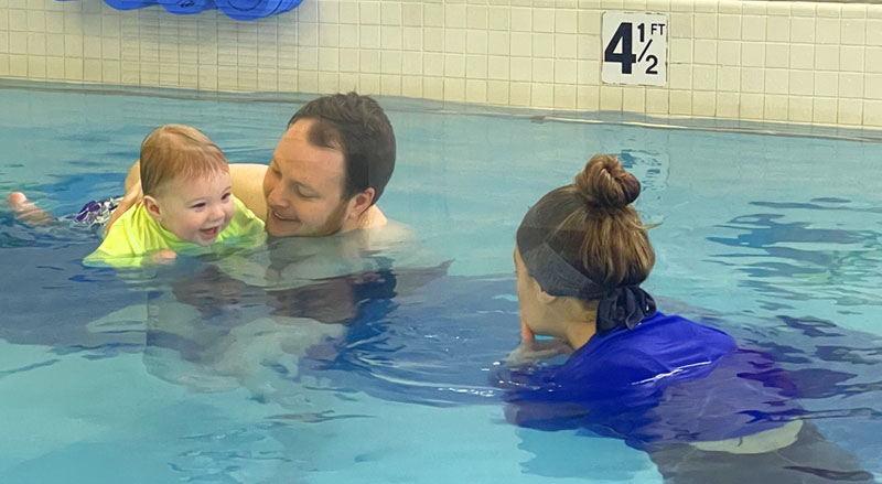 A smiling toddler in the pool with his father and swimming teacher