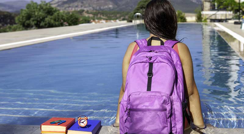 A young girl sitting on the edge of a pool with her school backpack on