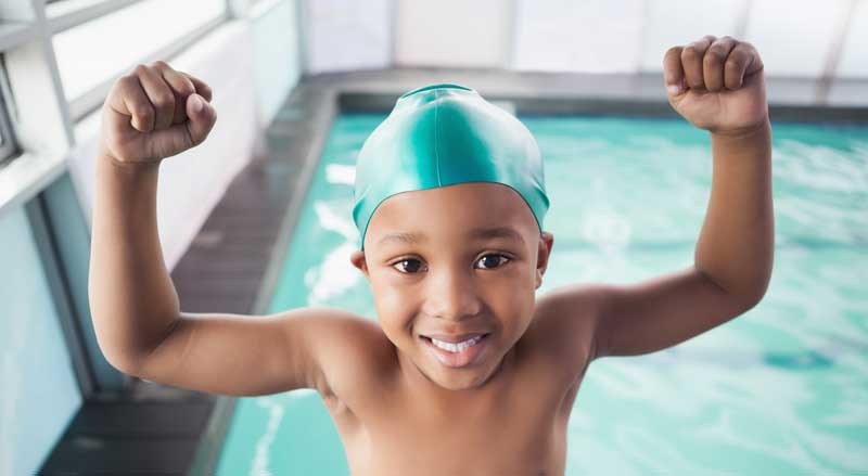 A young boy in a pool with his arms up displaying his strength and confidence. He looks proud of himself