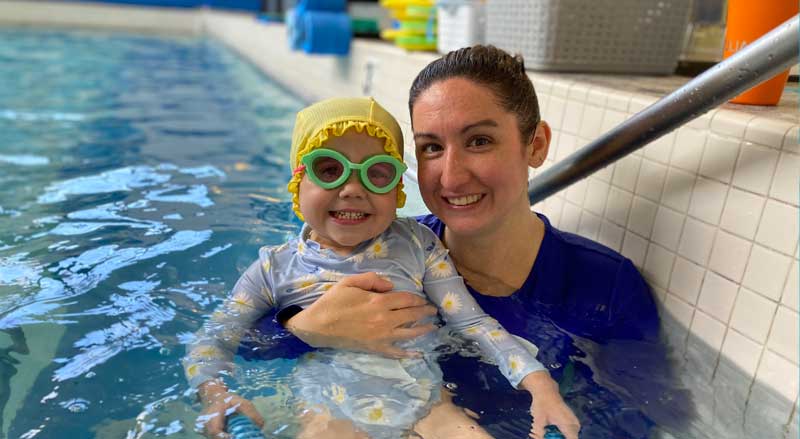 Njswim teacher holding a smiling toddler girl in the pool.