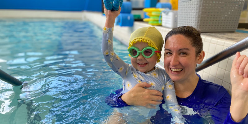 An Njswim swim teacher holding a smiling baby girl in the swimming pool
