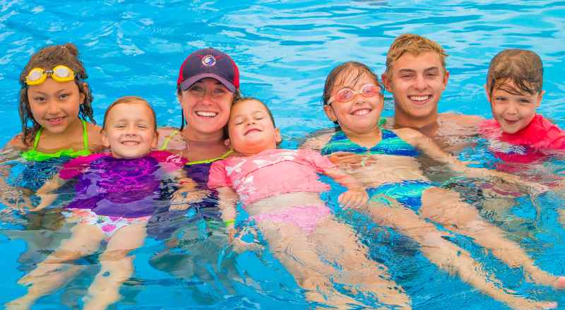 A joyful group of kids and two adults from the Njswim class are smiling and floating together in a swimming pool