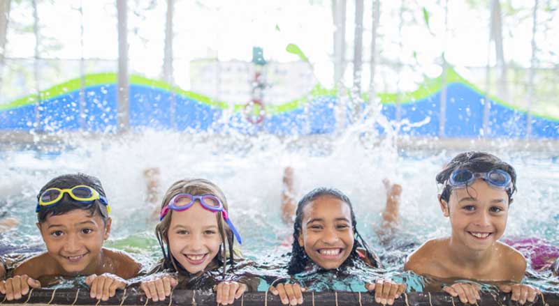 Smiling kids holding on to the edge of a pool practicing kicking in the water