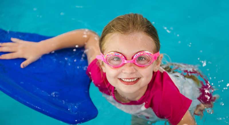 A smiling young girl wearing pink swim googles a pool.