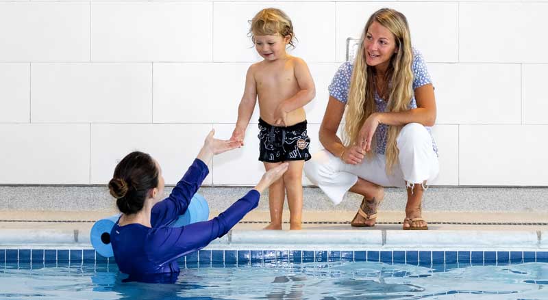 A swim teacher in a pool holding out her arms to young student standing next to parent on the pool deck