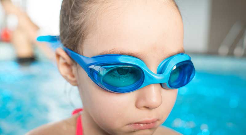 A young girl in swim goggles holding back tears
